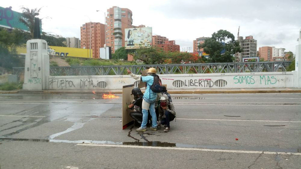 Die venezolanische Opposition im Straßenkampf auf der Brücke von Las Mercedes in Caracas geschützt durch Helme und ein Schutzschild am 7. Juni 2017. Auf der Wand Parolen wie „Weg mit der Diktatur!“, „Freiheit“ oder „Wir sind mehr.“